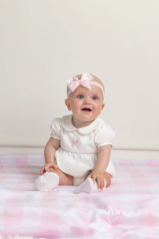 Baby in a white outfit with a pink headband sitting on a pink blanket.