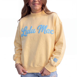 A woman wearing a pale yellow sweatshirt with 'Lulu Mac' embroidered in light blue across the chest, paired with light blue jeans against a plain white background.