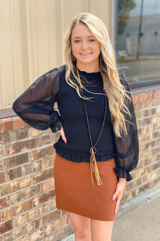 Woman standing in front of building in black top and toffee pencil skirt and black slides.